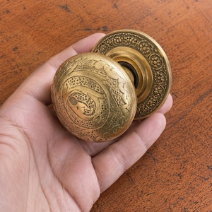 May include: A round brass door knob featuring intricate embossed floral and geometric patterns on the spherical handle and the circular backplate. The metal surface displays a warm, aged golden tone with detailed engravings across the entire exterior.