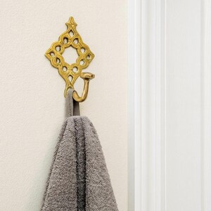 Pu&ograve; includere: Un gancio da parete in metallo dorato con una piastra posteriore decorata a forma di diamante, caratterizzata da un quadrato centrale aperto e dettagli a volute. Un asciugamano grigio testurizzato pende dal gancio curvo a parete.