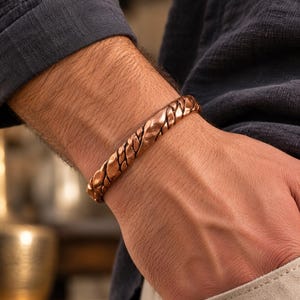 Peut inclure: Un bracelet manchette en cuivre présentant un design torsadé et tressé avec une finition martelée et texturée. Le métal possède un éclat métallique brun rougeâtre chaud et une forme arrondie continue qui entoure le poignet.