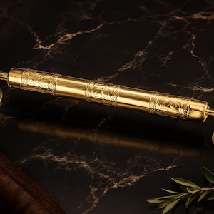 Könnte beinhalten: Ein polierter goldfarbener Türgriff aus Metall mit einer zylindrischen Stange, die zwei Abschnitte mit geprägten Blumenmustern aufweist. Der Griff wird von zwei geschwungenen Armen gehalten, die an runden Montageplatten mit Schraublöchern befestigt sind.