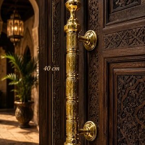 Puede incluir: Un tirador de puerta de latón pulido con intrincados patrones grabados y un diseño cilíndrico vertical. El tirador está montado en una puerta de madera oscura tallada y tiene una longitud total de 40 cm, según indican las marcas de medida.