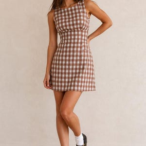 May include: A sleeveless mini dress featuring a brown and white checked pattern, a round neckline, and a fitted bodice that transitions into a slightly flared skirt. The dress is paired with white crew socks and black patent leather Mary Jane shoes.