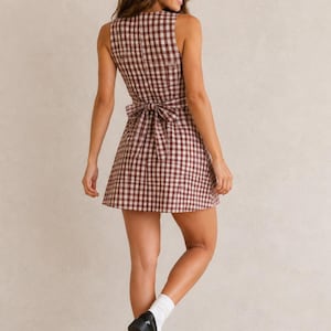 May include: A sleeveless mini dress featuring a brown and white gingham check pattern, a fitted bodice, and a tie-back waist detail. The dress has a straight neckline and a flared skirt that falls above the knee, paired with white socks and black shoes.