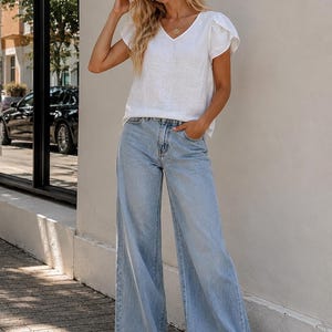 K&ouml;nnte beinhalten: Ein wei&szlig;es Oberteil mit V-Ausschnitt und kurzen, flatternden &Auml;rmeln, kombiniert mit einer hellblauen Jeans mit weitem Bein. Das Outfit umfasst ein wei&szlig;es Stoffstirnband, eine goldene Anh&auml;ngerkette und braune Sandalen mit offener Zehenpartie.
