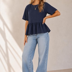 May include: A navy blue short-sleeved peplum top with a round neckline, paired with light-wash blue denim jeans that feature a wide-leg cut. The outfit is completed with brown thong sandals and a simple gold pendant necklace.
