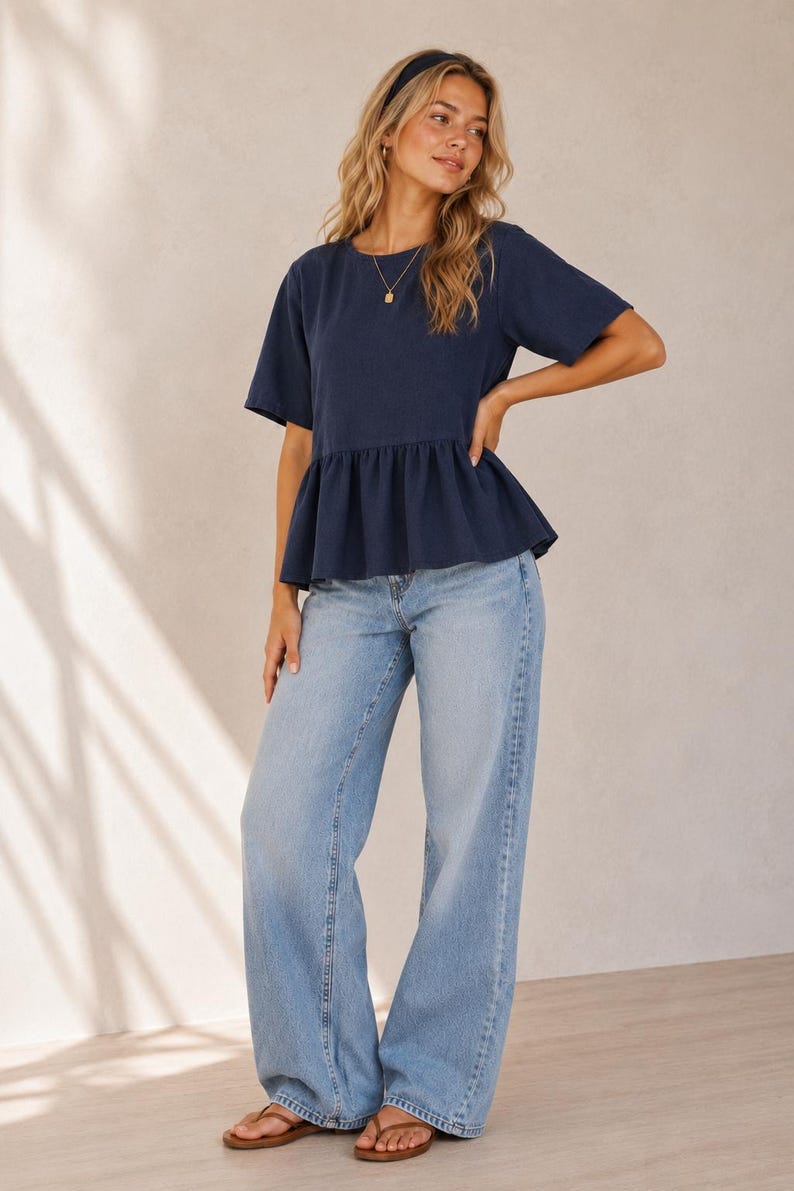 May include: A navy blue short-sleeved peplum top with a round neckline, paired with light-wash blue denim jeans that feature a wide-leg cut. The outfit is completed with brown thong sandals and a simple gold pendant necklace.