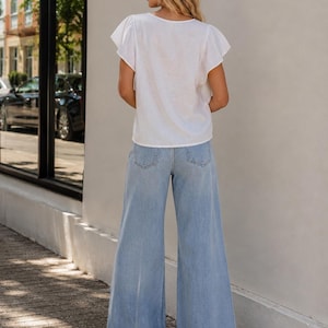 K&ouml;nnte beinhalten: Eine wei&szlig;e kurz&auml;rmelige Bluse mit Flatter&auml;rmeln und Rundhalsausschnitt, kombiniert mit einer hellblauen, weit geschnittenen Jeans. Die Bluse hat eine schlichte Textur, w&auml;hrend die Jeans ein klassisches F&uuml;nf-Taschen-Design mit sichtbaren Ges&auml;&szlig;taschen aufweist.