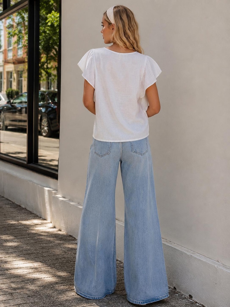 K&ouml;nnte beinhalten: Eine wei&szlig;e kurz&auml;rmelige Bluse mit Flatter&auml;rmeln und Rundhalsausschnitt, kombiniert mit einer hellblauen, weit geschnittenen Jeans. Die Bluse hat eine schlichte Textur, w&auml;hrend die Jeans ein klassisches F&uuml;nf-Taschen-Design mit sichtbaren Ges&auml;&szlig;taschen aufweist.