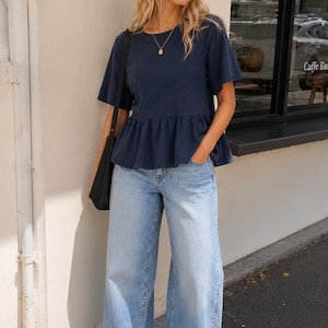 May include: A navy blue short-sleeved peplum top with a round neckline, paired with light-wash wide-leg denim jeans, a dark headband, a gold pendant necklace, a black shoulder bag, and brown flip-flop sandals.