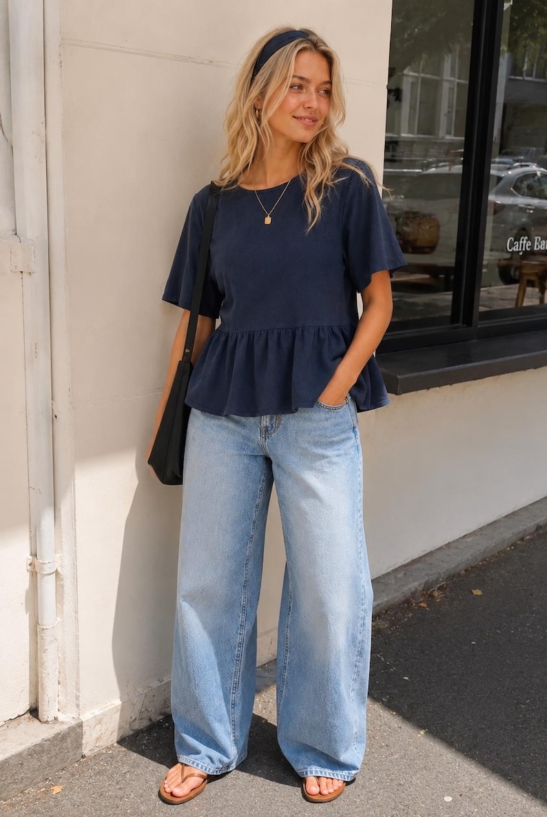 May include: A navy blue short-sleeved peplum top with a round neckline, paired with light-wash wide-leg denim jeans, a dark headband, a gold pendant necklace, a black shoulder bag, and brown flip-flop sandals.