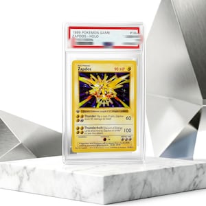 May include: A 1999 Pokemon Game Zapdos holographic trading card encased in a clear protective slab. The card features a yellow Zapdos illustration, 90 HP, and the attacks Thunder and Thunderbolt. The label at the top reads 1999 POKEMON GAME ZAPDOS.
