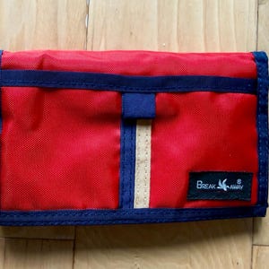 May include: A rectangular red nylon wallet with dark blue trim and a vertical beige stripe. The wallet features a dark blue horizontal strap across the top and a black rectangular patch in the bottom right corner with the text BREAK AWAY.