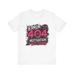 Puede incluir: Una camiseta blanca de manga corta con un diseño gráfico que incluye el texto ERROR 404 MOTIVATION NOT FOUND en rosa y blanco. El diseño presenta un icono de cara triste con ojos en forma de X, un triángulo de advertencia y salpicaduras negras.