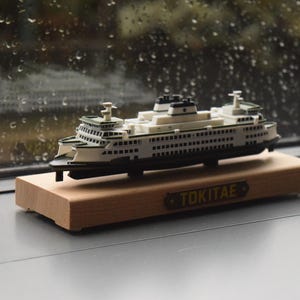 Könnte beinhalten: Ein Miniaturmodell einer Fähre mit weißem und dunkelgrünem Äußeren, mehreren Decks und Fenstern. Das Modell ist auf einem rechteckigen, hellen Holzsockel montiert, der eine kleine Plakette mit der schwarzen Aufschrift TOKITAE trägt.