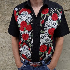 Peut inclure: Une chemise boutonnée à manches courtes noire avec un imprimé de crânes et de roses. La chemise a un col boutonné et une fermeture boutonnée.