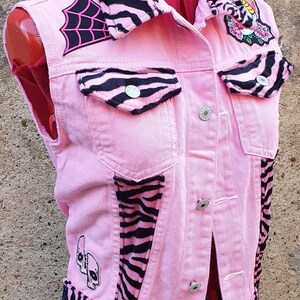 May include: Pink denim vest with black and white zebra print accents. Features patches including a spiderweb, skull, and floral design. The vest has silver buttons and is displayed on a mannequin.