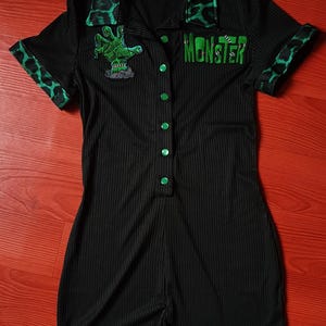 Può includere: Pagliaccetto nero con dettagli verdi e la scritta "MONSTER" in verde. Il colletto e i polsini presentano una stampa leopardata verde. Una toppa con la mano di un mostro verde è sul petto. Il pagliaccetto ha bottoni verdi.