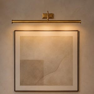 May include: A wall-mounted brass picture light illuminates a square framed print featuring beige and tan abstract shapes. Below, a floating dark wood console holds a textured ceramic vase, a stack of three books, and a small dark bowl.