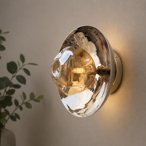 May include: A wall sconce featuring a polished silver-toned circular reflective backplate and a central, organic-shaped amber glass bulb. The light fixture emits a warm glow from the internal LED filament, creating a bright, mirrored surface effect.