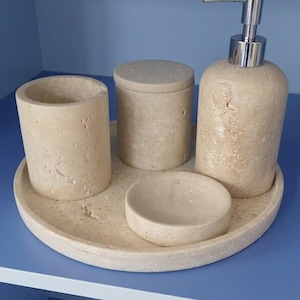 Peut inclure: Un ensemble d'accessoires de salle de bain en pierre naturelle beige et poreuse. Le set comprend un porte-brosse à dents cylindrique, un pot avec couvercle, un porte-savon et un distributeur de savon avec une pompe en métal argenté sur un plateau rond.
