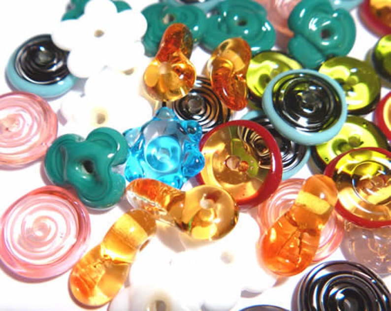 May include: A collection of colorful glass beads in various shapes and sizes. The beads include flower, spiral, and button designs in shades of white, blue, green, orange, and red. Some beads have a glossy finish, while others are translucent.