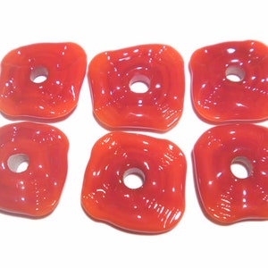May include: Six orange-red, square-shaped glass beads with a hole in the center. The beads have rounded edges and a glossy finish. They are arranged on a white surface, showcasing their color and shape.