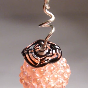 May include: A handmade earring featuring a cluster of peach-colored beads topped with a black and clear glass accent. The earring hangs from a silver-toned wire with a spiral design.