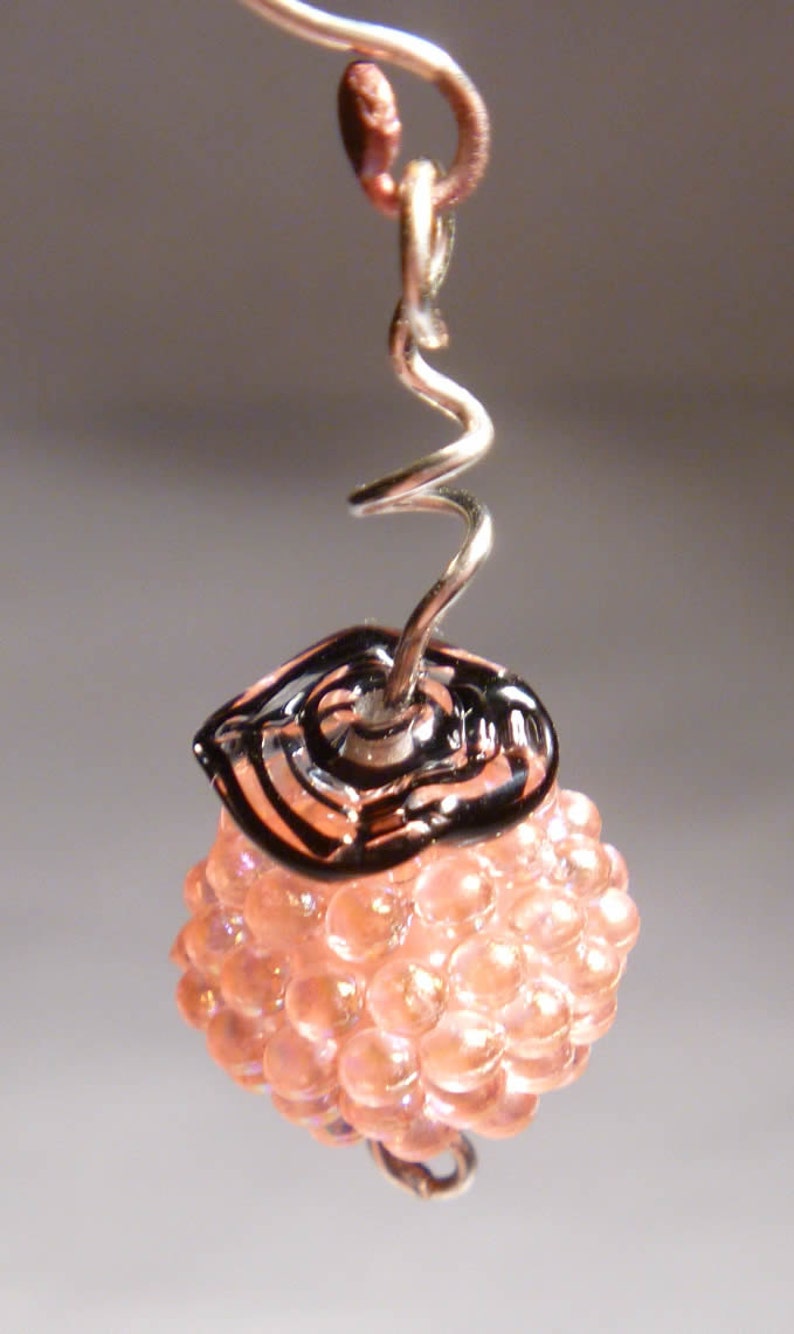 May include: A handmade earring featuring a cluster of peach-colored beads topped with a black and clear glass accent. The earring hangs from a silver-toned wire with a spiral design.