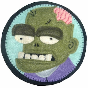 May include: A circular felt patch featuring a green zombie face with white teeth and a pink exposed brain. The zombie has dark brown eyes and is set against a light blue background, framed by a black border with white stitching. The zombie wears a purple shirt.