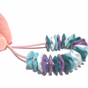 May include: A colorful bracelet featuring a series of flattened, irregular beads in shades of teal, white, and purple. The beads are strung on a pink cord, creating a unique and eye-catching accessory.