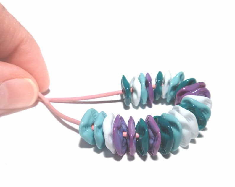 May include: A colorful bracelet featuring a series of flattened, irregular beads in shades of teal, white, and purple. The beads are strung on a pink cord, creating a unique and eye-catching accessory.
