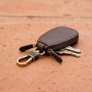 May include: A dark brown, pebble-textured leather key case with a zipper closure. The case features a brass-toned metal lobster clasp attached to a leather tab and contains several silver-toned metal keys protruding from the zippered opening.