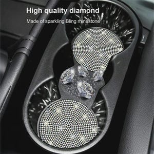 May include: A black car cup holder insert featuring two circular pads covered in sparkling clear rhinestones, connected by a central piece decorated with four large, faceted diamond-shaped crystals. Text reads: High quality diamond. Made of sparkling Bling rhinestone.