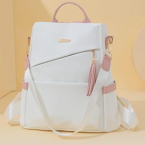 May include: A white backpack featuring light pink accents on the top handle, side straps, and base. The bag includes a diagonal front zipper pocket adorned with a hanging pink tassel, a gold-toned metal emblem, and adjustable white shoulder straps.