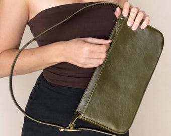 May include: A rectangular olive green shoulder bag with a smooth finish, featuring a single thin strap, a top zipper closure, and gold-toned metal hardware. The bag has a structured, elongated shape and a minimalist design with clean stitching.
