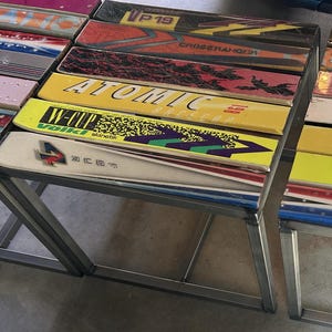 May include: A table constructed from repurposed vintage skis featuring various colorful designs and brand logos including Atomic, Volkl, and Crossranger. The tabletop is composed of multiple parallel skis with a metal frame base supporting the structure.