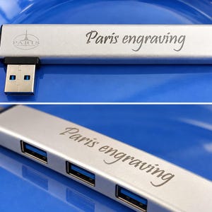 May include: A silver-colored USB hub featuring three blue-accented ports and a black connector. The device is engraved with the text "Paris engraving" and a small Eiffel Tower icon. The metallic surface has a smooth finish and a rectangular shape.