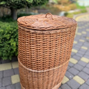 May include: A tall, oval-shaped laundry hamper crafted from woven brown wicker. The hamper features a matching removable lid with a small, centered loop handle on top. The exterior displays a consistent, tight weave pattern throughout the body.