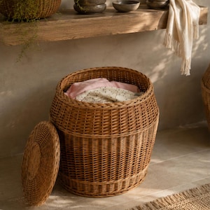 May include: A round, barrel-shaped wicker laundry basket with a woven texture and a matching removable lid. The basket is filled with folded light-colored textiles, including a pink garment and a white fabric with small dark dots.