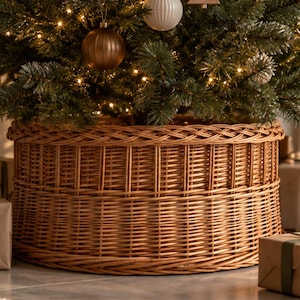 May include: A round, woven wicker tree collar with a natural light brown finish. The base features a sturdy, cylindrical design with a braided top rim and vertical reed detailing, designed to conceal the base of a Christmas tree.