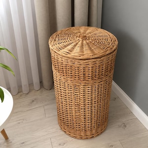 Puede incluir: Un cesto para la ropa alto y cilíndrico fabricado en mimbre trenzado de color marrón claro. El cesto cuenta con una tapa circular a juego con un patrón de tejido radiante y un diseño de tejido vertical en los laterales, creando una unidad de almacenamiento.