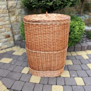 May include: A tall, oval-shaped laundry hamper crafted from light brown woven wicker. The hamper features a matching removable lid with a small central handle and a reinforced horizontal band running around the middle of the vertical body structure.