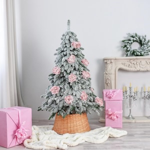 Peut inclure: Un sapin de Noël artificiel floqué décoré de fleurs de roses rose pâle, posé dans une base en panier d'osier tressé. L'arbre est entouré de boîtes cadeaux rose uni nouées avec des rubans et d'un tapis en tissu blanc texturé.