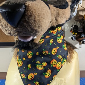 May include: A black bandana with a Halloween-themed print of orange jack-o'-lantern faces on a black background with yellow stars. The bandana is tied around the neck of a furry brown and black animal head.