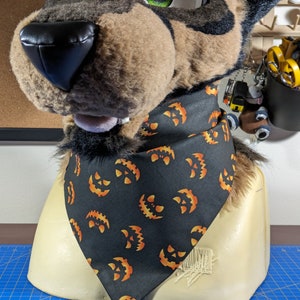 May include: A black Halloween-themed bandana featuring a repeating pattern of orange jack-o'-lantern faces. The bandana is triangular and is worn around the neck of a furry animal head. The head is brown and tan with green accents.