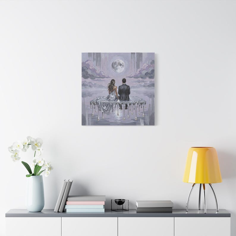 A small, framed oil painting of a couple on a dripping silver platform under a full moon hangs on a white wall above a gray console table with books and a yellow lamp.