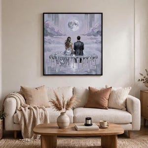 A framed oil painting of a couple on a dripping silver platform under a full moon hangs above a white sofa with pillows, a coffee table, and a potted plant.
