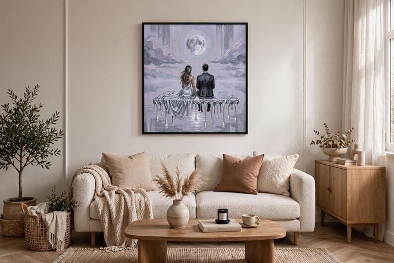 A framed oil painting of a couple on a dripping silver platform under a full moon hangs above a white sofa with pillows, a coffee table, and a potted plant.