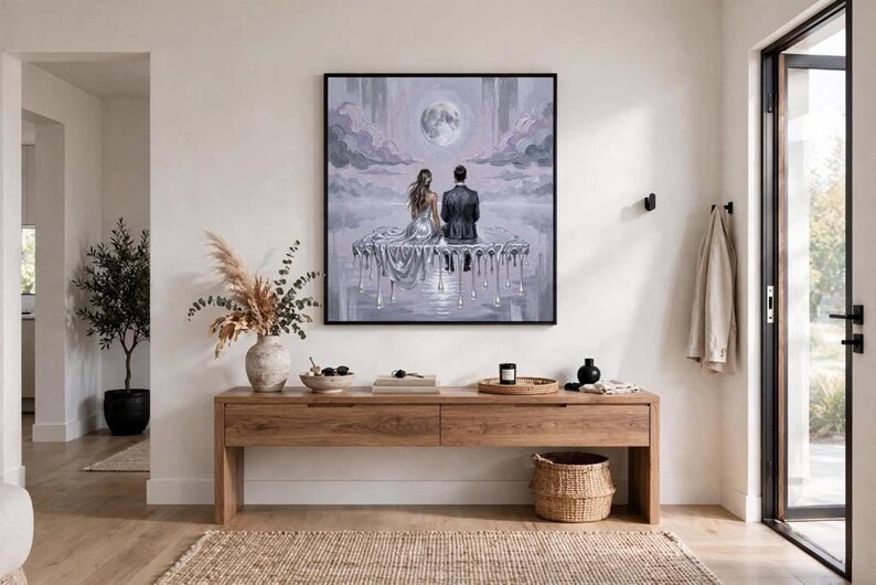 A large, framed oil painting of a couple on a dripping silver platform under a full moon hangs above a wooden console table in a minimalist room with a potted plant.