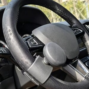 Può includere: Un coprivolante nero con texture traforata e un pomello per volante nero solido fissato al bordo. La copertura presenta un sottile motivo a rombi impresso nel materiale, mentre il pomello è di forma rotonda e liscia.